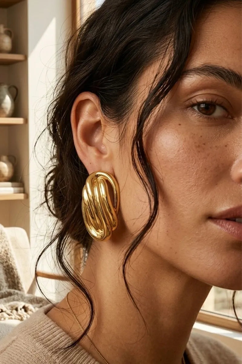 A close-up photograph of a woman wearing large, polished gold knot earrings, with her dark curly hair partially covering one ear. The earrings have an intertwined, chunky design.
