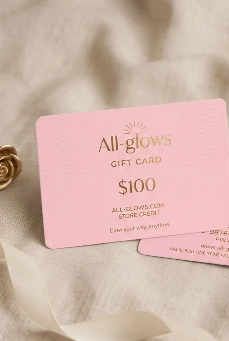 All-glows $100 fashion jewelry gift card in elegant pink with gold foil lettering, styled alongside a metallic golden rose and silk ribbon on a soft linen fabric background.