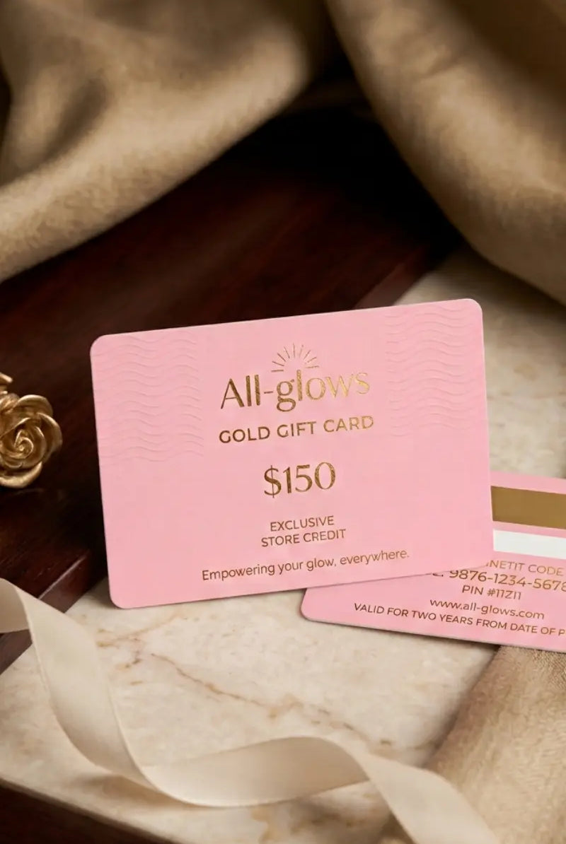 All-glows $150 Gold Gift Card featuring embossed pink card design, displayed on a luxurious dark wood and white marble surface with textured drapes and a gold rose accent.