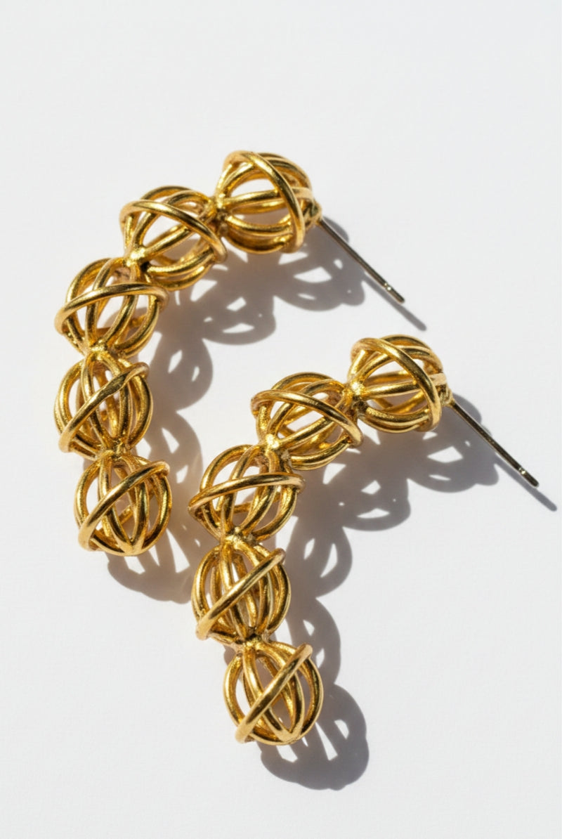 A pairs of architectural 18K gold-plated wire-woven hoop earrings placed on a crisp white surface under direct sunlight, displaying bold geometric overlapping lines and sharp, hollow-core structure shadows.