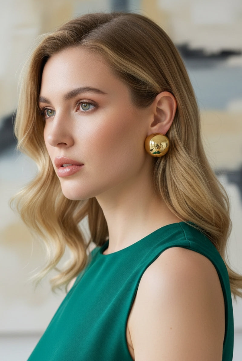 Profile view of a blonde model wearing a green top and All-Glows contemporary brass jewelry. The oversized gold dome studs deliver a bold runway-inspired look while maintaining a comfortable, lightweight fit for all-day wear.