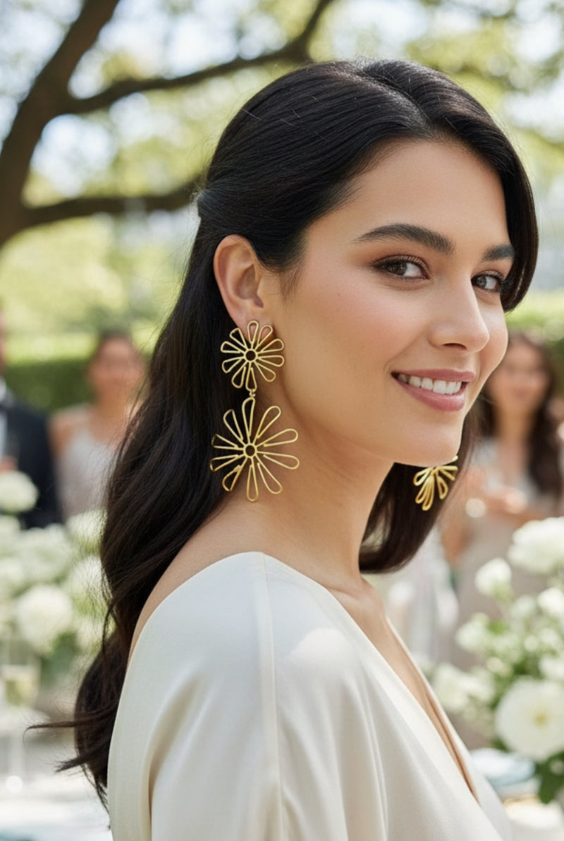 Smiling woman wearing long gold double daisy floral earrings at an outdoor garden party.