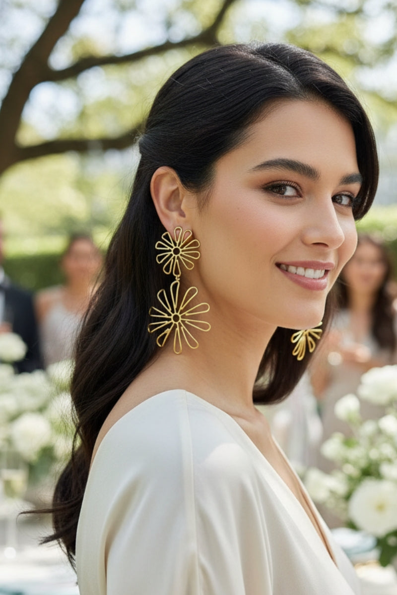 Smiling woman wearing long gold double daisy floral earrings at an outdoor garden party.