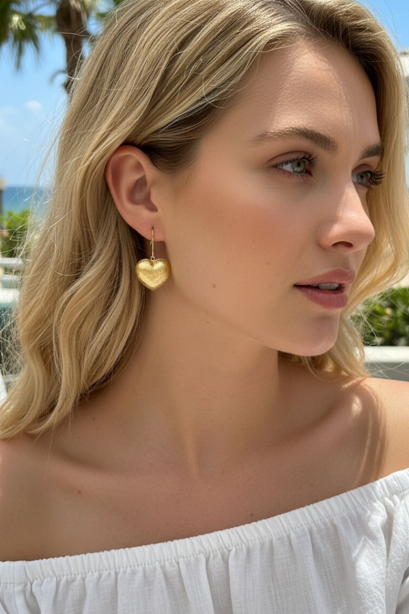 A woman with wavy blonde hair wearing textured gold heart-shaped drop earrings against a beautiful blue ocean backdrop. Perfect lightweight summer jewelry.