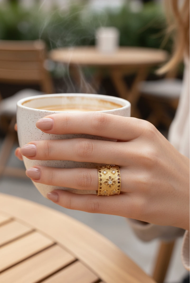 A model with manicured beige nails holds a latte cup in a cafe, showcasing the intricate brushed gold filigree pattern of the All Glows Ash ring.