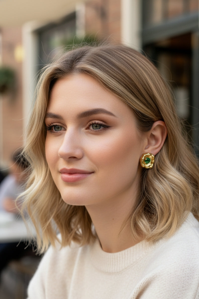 Natural outdoor lifestyle portrait of a blonde woman with a shoulder-length bob wearing contemporary indie designer organic gold earrings with green crystal inserts. Minimalist capsule wardrobe jewelry styling for autumn and winter.
