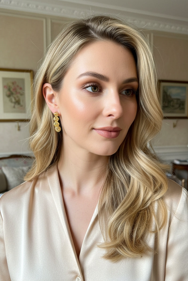 A warm-toned profile shot of a woman with honey-blonde hair dressed in a luxurious champagne silk blouse, accessorized with the classic 4-orb gold statement studs to illustrate an effortless French chic styling ideal for daily luxury.