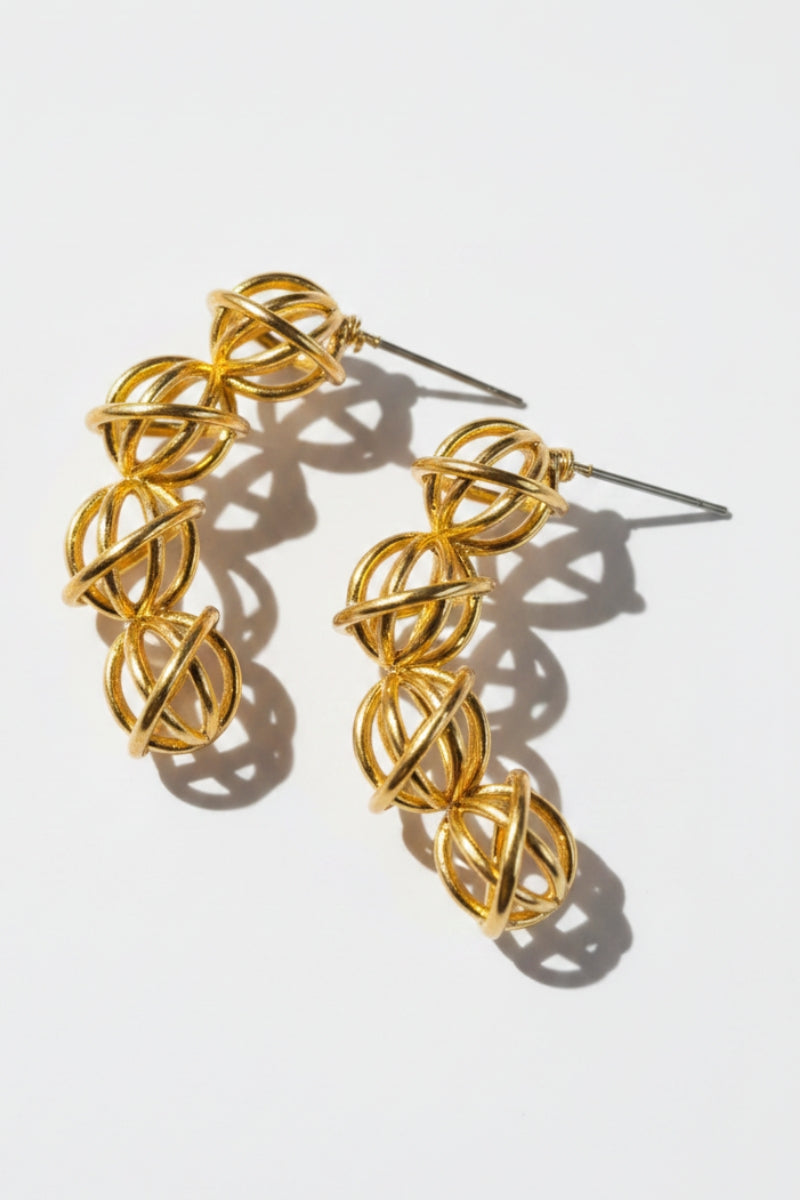 Two hollow gold ball statement studs layout flat under strong daylight, casting dramatic geometric shadows that emphasize the high-shine 18K gold plating and delicate metallic wire craftsmanship.