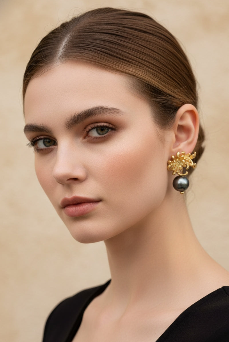 Symmetrical portrait of a woman with a clean low bun wearing artisanal 18K gold-plated orchid flower earrings with swinging black pearls. Organic modernism style jewelry shot against a warm beige textured wall.