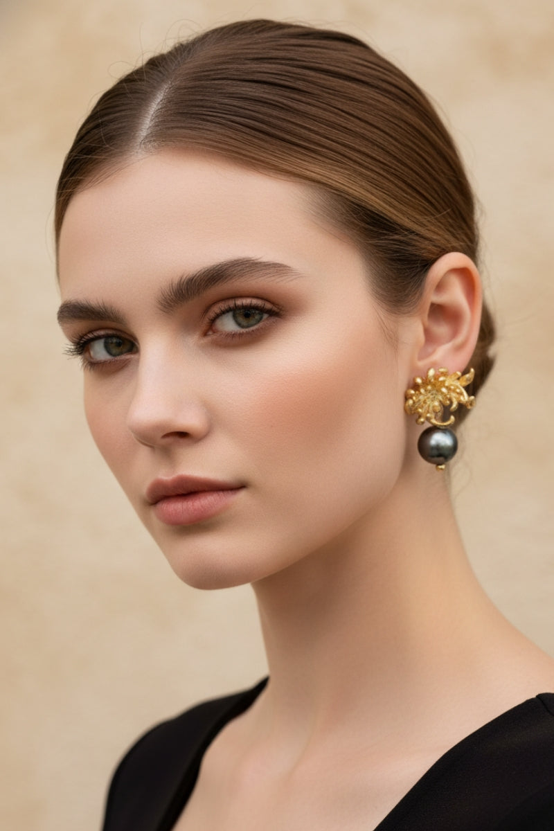 Symmetrical portrait of a woman with a clean low bun wearing artisanal 18K gold-plated orchid flower earrings with swinging black pearls. Organic modernism style jewelry shot against a warm beige textured wall.