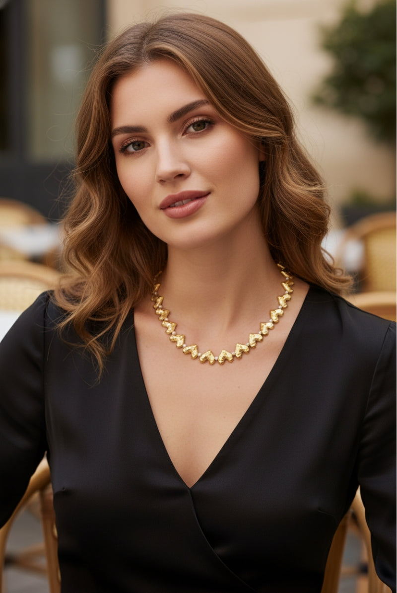 Beautiful woman wearing the Jupiter gold necklace with interlocking heart charms and small pearl accents, paired with a black V-neck dress in a Parisian-style cafe. High-angle portrait.