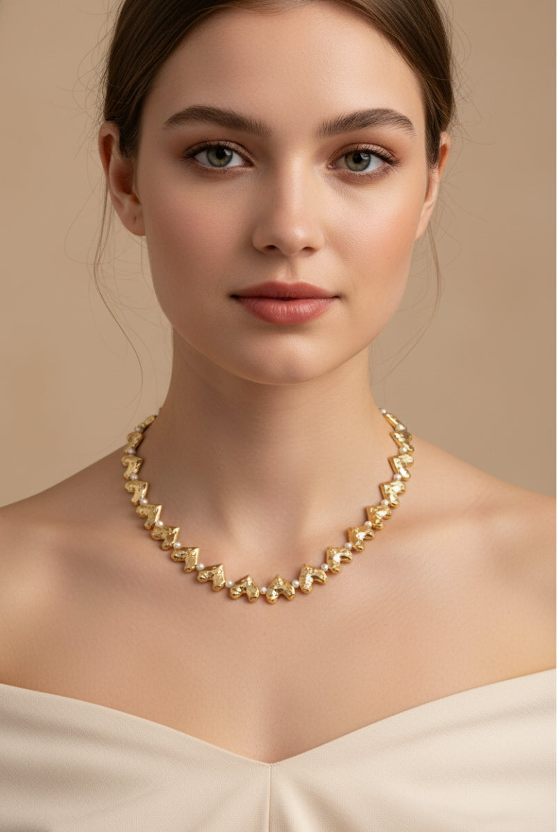 Detail view of the Jupiter repeating heart design necklace, crafted with gold charms and interspersed small round white pearls. Worn on an off-the-shoulder cream top. Flat lay alternative.