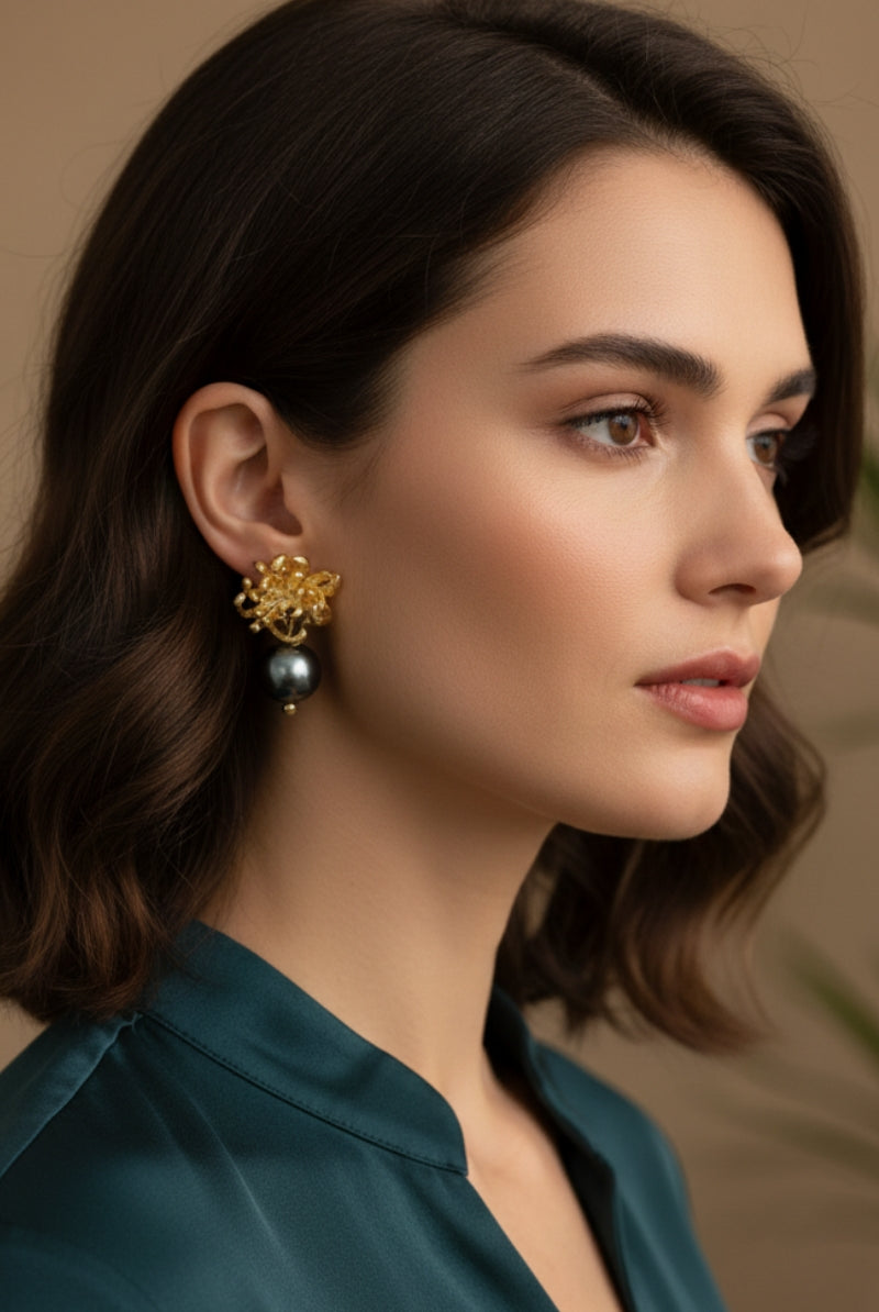 A brunette woman with wavy hair wearing heavy-textured gold botanical metal earrings with dark tahitian style pearls. High-contrast styling capturing bold mob wife aesthetic accessories with an emerald green satin shirt.