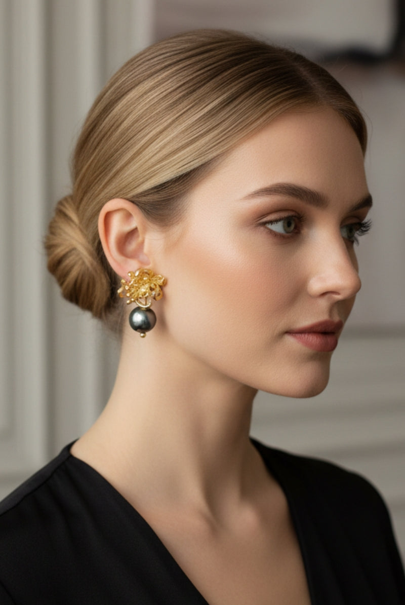 Three-quarter view of a woman with low tied hair wearing boutique quality textured gold flower earrings with a light-catching dark pearl. Minimalist modern vintage capsule collection jewelry demonstration.