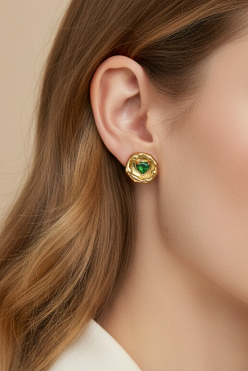 Super close-up of a woman with honey blonde hair wearing an 18K gold-plated molten metal stud earring with a heart-shaped green emerald crystal center. Clean minimalist jewelry shot focusing on the liquid metal textured edge.