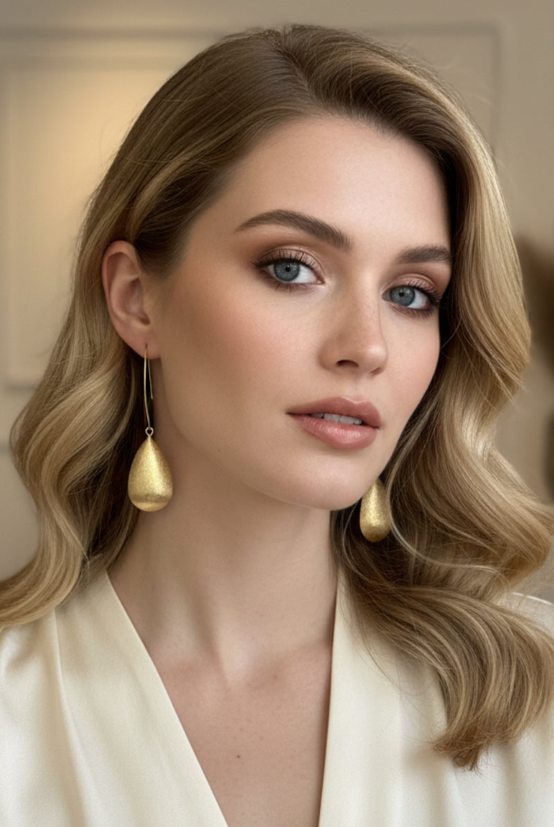 Full-face portrait of a woman with elegant wavy blonde hair and soft glamorous makeup, wearing the long Queen earrings. The matte-finish gold teardrop pendants hang perfectly, framing her face. Worn with a white silk v-neck.