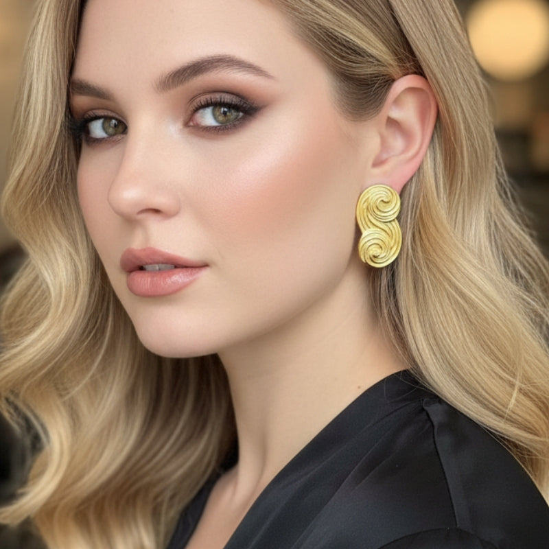 A close-up portrait of a model with wavy blonde hair showcasing All Glows Seraphina earrings, which feature a unique, sculptural gold swirl design, for an upscale evening look.