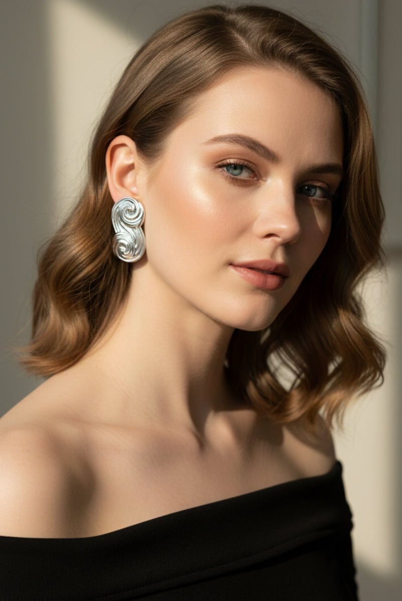 A detailed shot of the Seraphina Sculptural Swirl Earrings worn with a black off-the-shoulder top. These silver statement studs add a contemporary, artistic touch to a classic minimalist outfit.