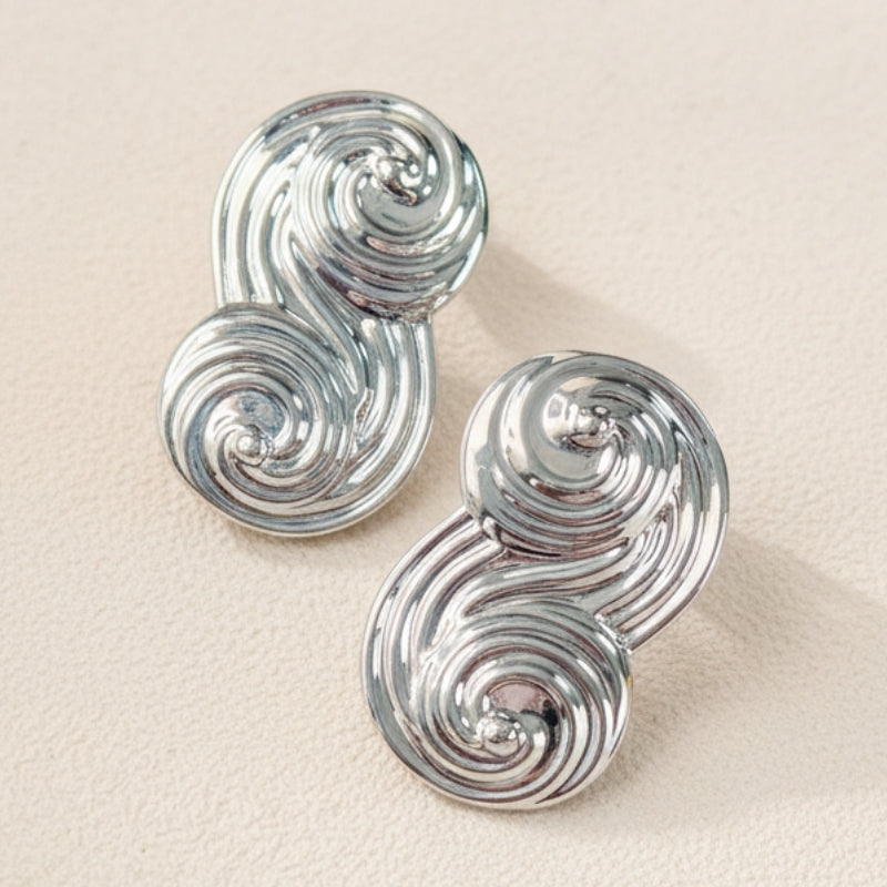 A pair of Seraphina Sculptural Swirl Earrings displayed on a textured beige background. The photo shows the intricate grooved texture and mirror-like silver finish of these artistic statement earrings.