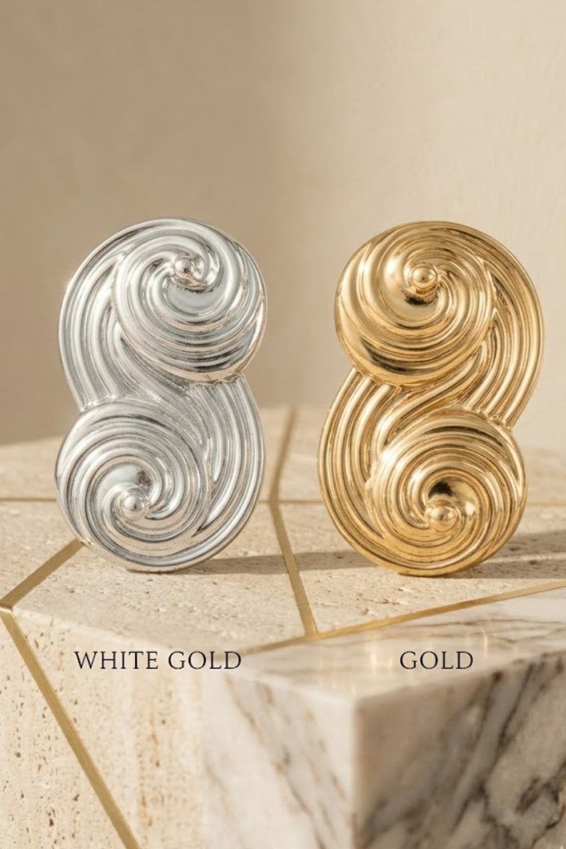 A high-end professional comparison shot of Seraphina's signature sculptural S-shaped swirl stud earrings. The image is split vertically to showcase two premium finishes: the left side features the radiant White Gold / Platinum finish on a light grey marble base, and the right side features the rich Yellow Gold finish on a warm beige travertine stone. Above each earring, the text clearly identifies 'WHITE GOLD' and 'YELLOW GOLD' in an elegant serif font. The design highlights the intricate, flowing architect