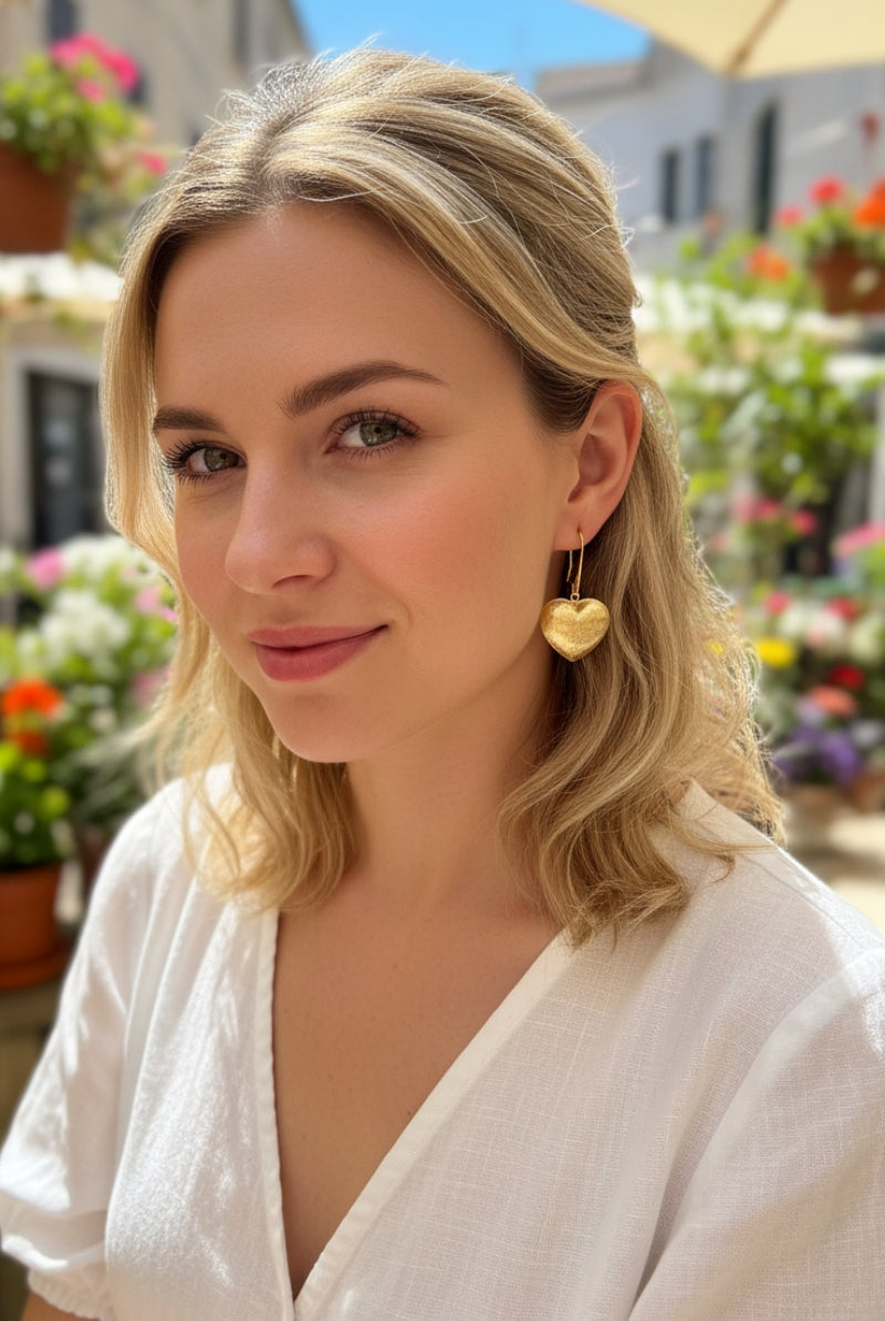 Close-up of a woman in a white linen top wearing heart-shaped gold-plated earrings in a sun-drenched flower garden. Sophisticated everyday luxury.