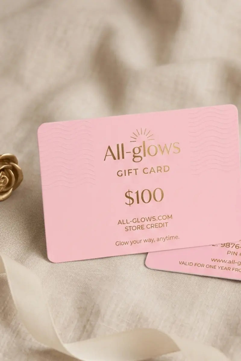 All-glows $100 fashion jewelry gift card in elegant pink with gold foil lettering, styled alongside a metallic golden rose and silk ribbon on a soft linen fabric background.