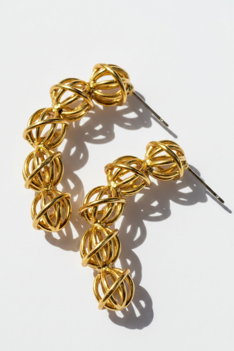 A pairs of architectural 18K gold-plated wire-woven hoop earrings placed on a crisp white surface under direct sunlight, displaying bold geometric overlapping lines and sharp, hollow-core structure shadows.