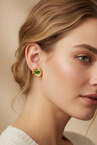 Close-up studio portrait of a soft smiling woman with swept-back hair showcasing a sculptural gold stud earring with a central green crystal. Perfect everyday luxury accessory inspiration for professional women seeking tarnish-resistant jewelry.