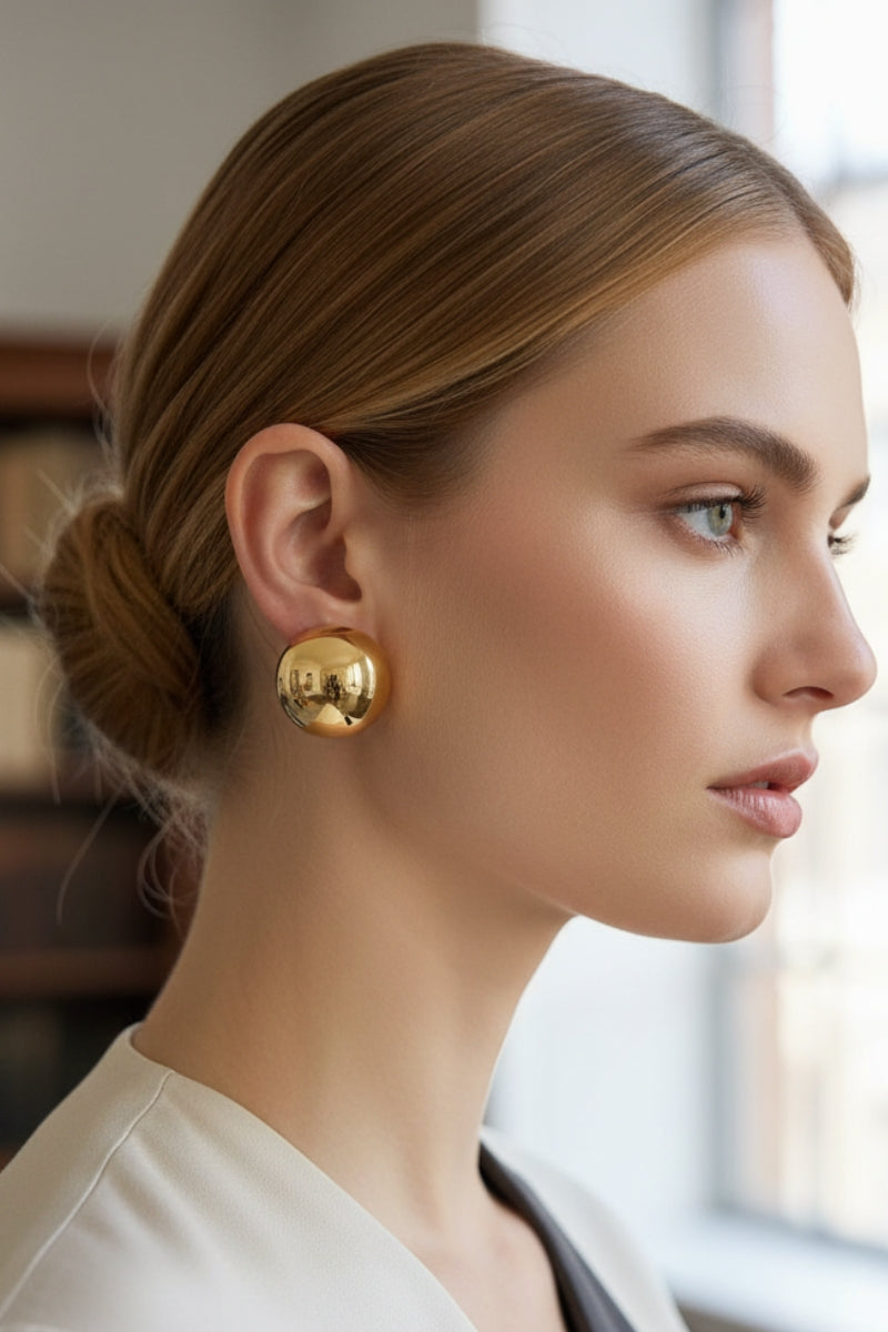 Side profile of a woman with her hair in a sleek bun, wearing All-Glows Aura Sculpted Dome Studs, which are large, chunky 18K gold-plated sphere earrings reflecting a brilliant mirror finish.