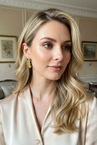 A warm-toned profile shot of a woman with honey-blonde hair dressed in a luxurious champagne silk blouse, accessorized with the classic 4-orb gold statement studs to illustrate an effortless French chic styling ideal for daily luxury.