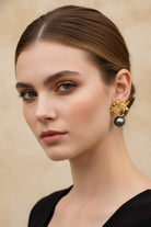 Symmetrical portrait of a woman with a clean low bun wearing artisanal 18K gold-plated orchid flower earrings with swinging black pearls. Organic modernism style jewelry shot against a warm beige textured wall.
