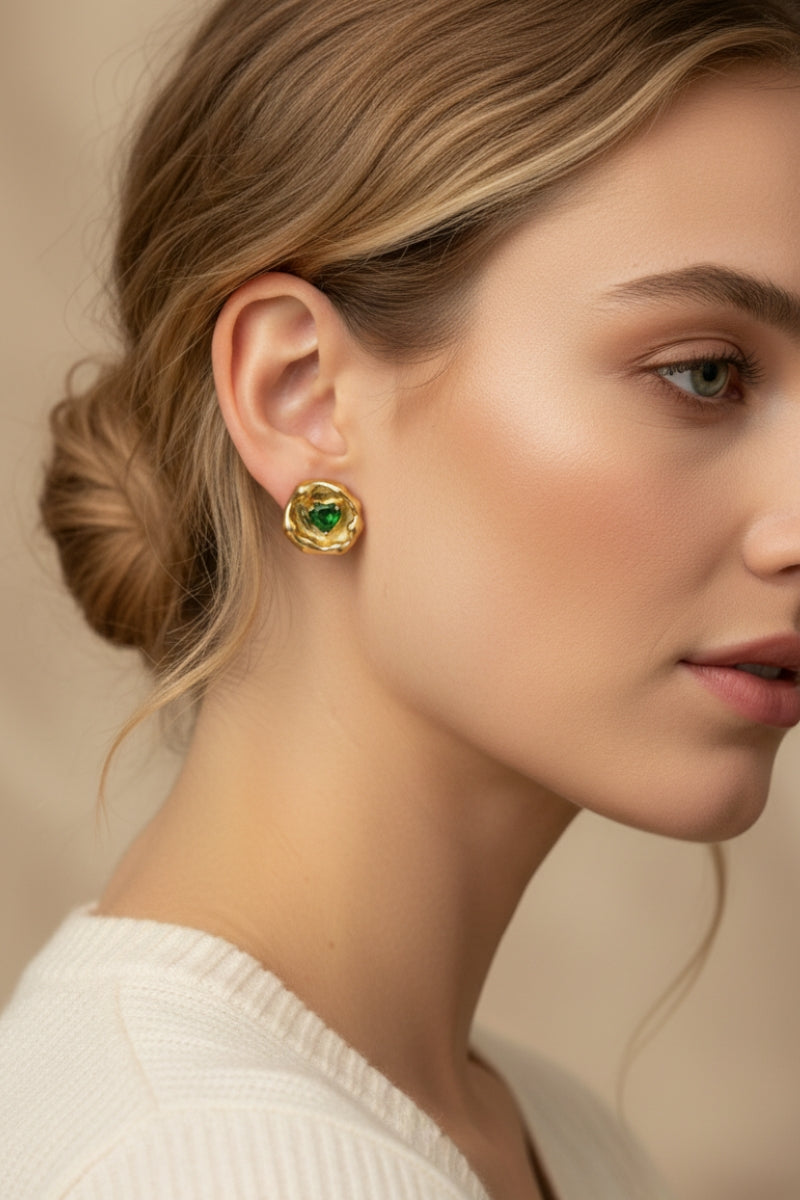 Side profile photo of a blonde woman with an elegant low bun wearing textured crumpled gold molten studs featuring a vibrant green gemstone. Demonstrating how to style quiet luxury artisan jewelry with a casual white rib-knit sweater.