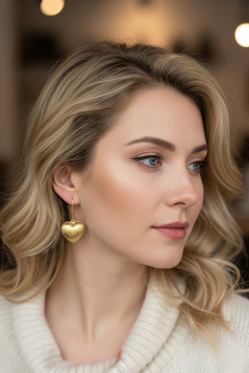 A woman wearing a cream turtleneck sweater paired with brushed gold heart earrings. Detailed view of the handcrafted metal texture in warm indoor lighting.