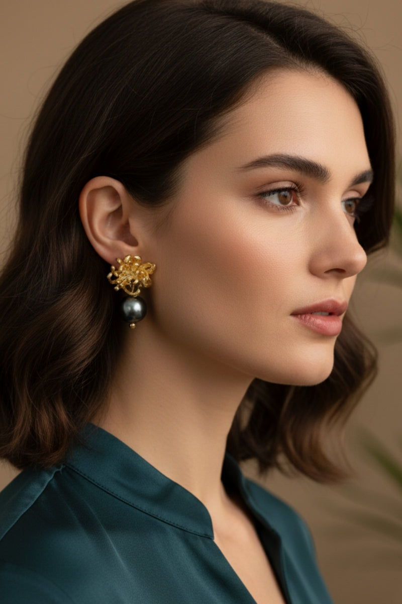 A brunette woman with wavy hair wearing heavy-textured gold botanical metal earrings with dark tahitian style pearls. High-contrast styling capturing bold mob wife aesthetic accessories with an emerald green satin shirt.