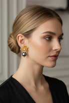 Three-quarter view of a woman with low tied hair wearing boutique quality textured gold flower earrings with a light-catching dark pearl. Minimalist modern vintage capsule collection jewelry demonstration.