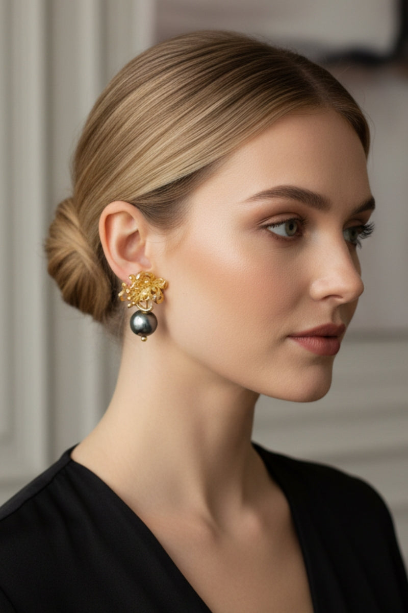 Three-quarter view of a woman with low tied hair wearing boutique quality textured gold flower earrings with a light-catching dark pearl. Minimalist modern vintage capsule collection jewelry demonstration.