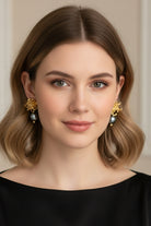 Front view portrait of a woman wearing a matching pair of nickel-free statement drops featuring detailed gold chrysanthemum floral bases and symmetrical tahitian-style black pearls, ideal for timeless formal styling.