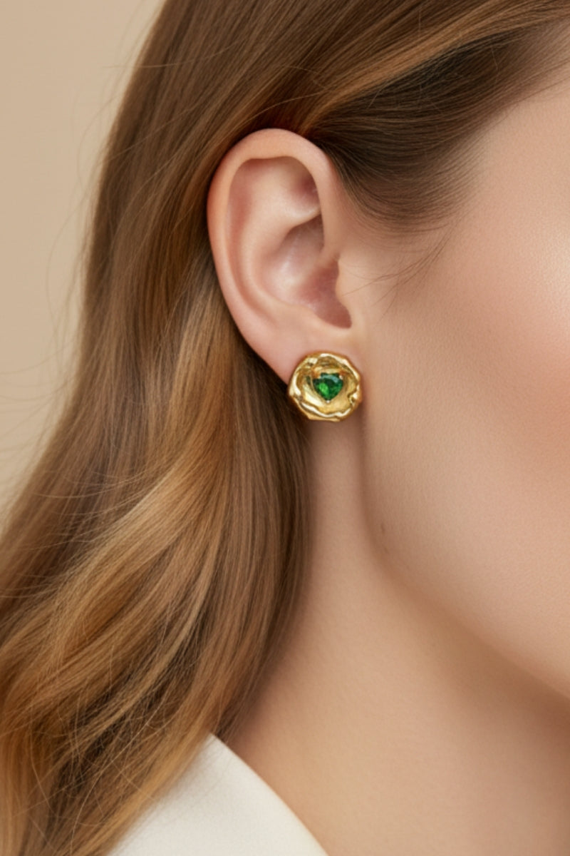 Super close-up of a woman with honey blonde hair wearing an 18K gold-plated molten metal stud earring with a heart-shaped green emerald crystal center. Clean minimalist jewelry shot focusing on the liquid metal textured edge.