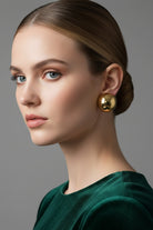 An elegant woman in an emerald green velvet dress accessorized with Aura Sculpted Dome Studs, demonstrating how these premium 18K gold plated chunky sphere earrings add an effortless touch of geometric luxury to evening wear.