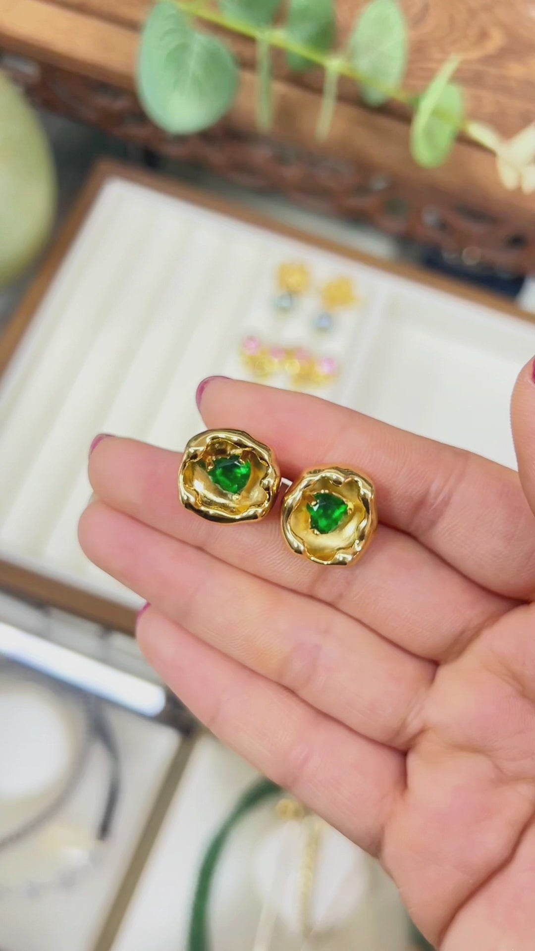 A high-definition close-up video of a hand holding and tilting a pair of 18K gold-plated molten heart stud earrings to showcase the rich liquid metal aesthetic and irregular lava texture. The emerald green gemstone in the center sparkles as it catches the light, demonstrating how to choose hypoallergenic sterling silver post earrings for sensitive ears and capturing quiet luxury jewelry inspiration for everyday modern vintage capsule wardrobes.