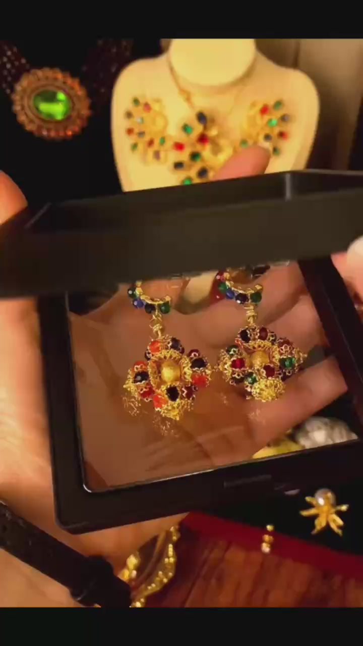 A vibrant close-up video showcase of tarnish-resistant gold dangles, featuring a bold, openwork cross-shaped setting filled with round multi-color gemstones, including red, green, blue, yellow, and pastel tones. A hand holds and rotates the earrings, suitable for a sophisticated evening party look.