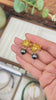 A close-up video of a hand holding and showcasing a pair of 18K gold-plated molten orchid flower earrings with swinging dark obsidian-toned pearl drops over a luxury jewelry display box. The video highlights the intricate handmade wax-carving texture of the botanical metalwork and the lustrous reflection of the dark pearls, answering how to find high-quality, tarnish-resistant statement jewelry and unique birthday gift ideas for her.