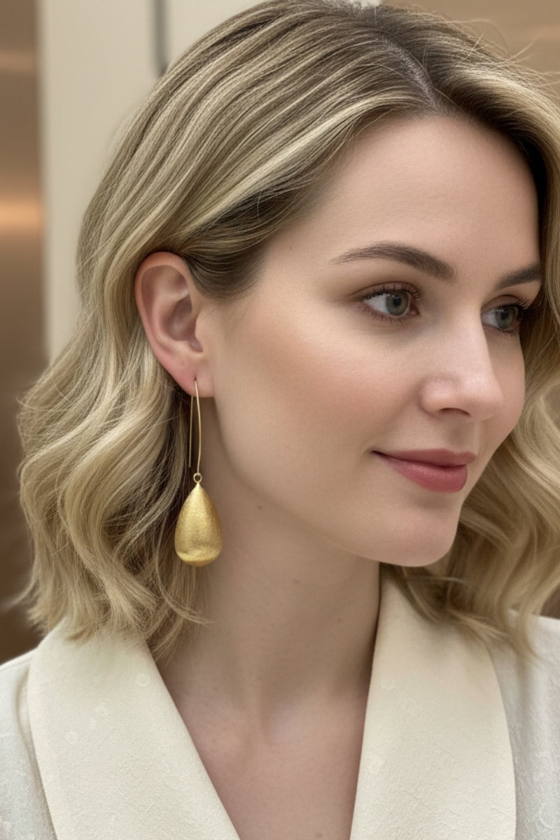 Side profile view of a smiling woman with wavy blonde hair wearing the long Queen earrings, which feature a distinct matte-brushed gold teardrop pendant on a fine hook. Styled with an off-white blouse.