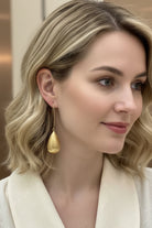 Side profile view of a smiling woman with wavy blonde hair wearing the long Queen earrings, which feature a distinct matte-brushed gold teardrop pendant on a fine hook. Styled with an off-white blouse.