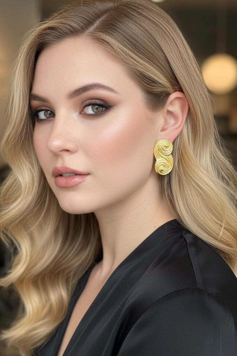 A close-up portrait of a model with wavy blonde hair showcasing All Glows Seraphina earrings, which feature a unique, sculptural gold swirl design, for an upscale evening look.