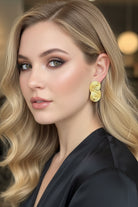 A close-up portrait of a model with wavy blonde hair showcasing All Glows Seraphina earrings, which feature a unique, sculptural gold swirl design, for an upscale evening look.