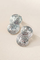 A pair of Seraphina Sculptural Swirl Earrings displayed on a textured beige background. The photo shows the intricate grooved texture and mirror-like silver finish of these artistic statement earrings.