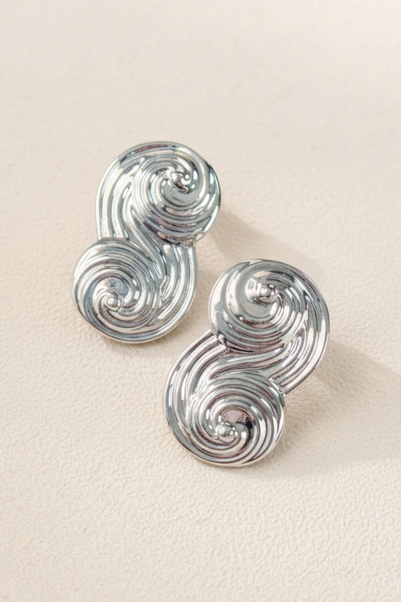A pair of Seraphina Sculptural Swirl Earrings displayed on a textured beige background. The photo shows the intricate grooved texture and mirror-like silver finish of these artistic statement earrings.