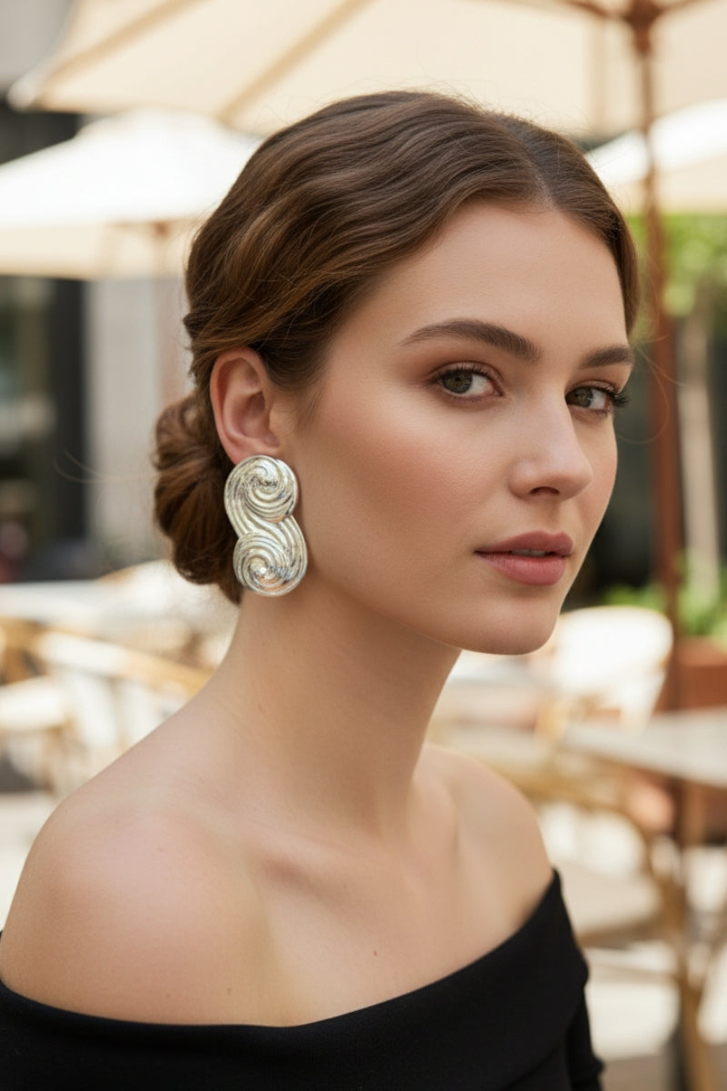 Close-up of a woman with an elegant updo hairstyle wearing Seraphina Sculptural Swirl Earrings in silver. These chunky high-shine earrings feature a distinctive double-spiral design, perfect for sophisticated evening wear.