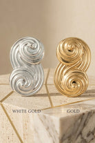 A high-end professional comparison shot of Seraphina's signature sculptural S-shaped swirl stud earrings. The image is split vertically to showcase two premium finishes: the left side features the radiant White Gold / Platinum finish on a light grey marble base, and the right side features the rich Yellow Gold finish on a warm beige travertine stone. Above each earring, the text clearly identifies 'WHITE GOLD' and 'YELLOW GOLD' in an elegant serif font. The design highlights the intricate, flowing architect
